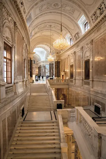 Eine prächtige Treppe in einem historischen Gebäude mit einer hohen Decke und kunstvoll gestalteten Wänden. Ein Kronleuchter hängt von der Decke, während große Fenster Tageslicht hereinlassen. Die Stufen führen hinunter zu einem offenen Bereich, der mit Stühlen und Tischen ausgestattet ist.