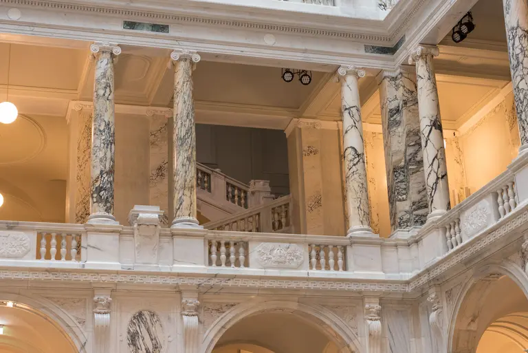 Eine Architekturaufnahme zeigt einen Innenraum mit eleganten Säulen aus Marmor und einer Treppe im Hintergrund. Die Wände sind mit historischen Details verziert, und die Beleuchtung sorgt für eine helle, einladende Atmosphäre.